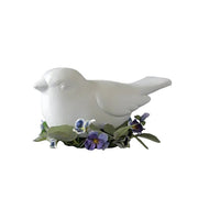 White bird figurine in classical style, surrounded by ornate violet flowers and green leaves, evoking old world European charm.