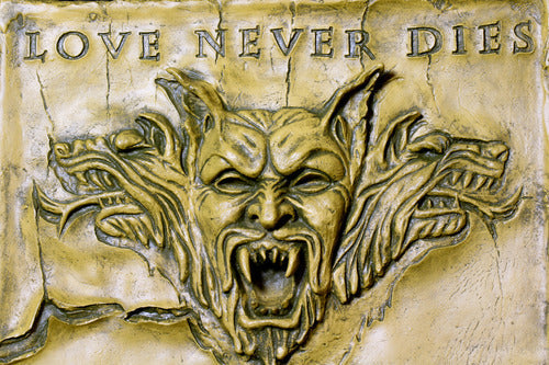 Baroque-style ornate plaque featuring a fierce, snarling gargoyle face with three wolf heads and "Love Never Dies" inscription above.