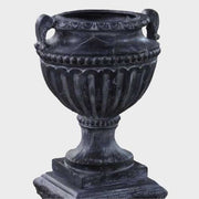Ornate classical urn with fluted detailing and handles, set atop a square pedestal with intricate old world European reliefs. Side Angle
