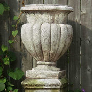 Ornate classical urn planter with fluted Baroque detailing on a square pedestal, evoking old world European garden style. Side Angle